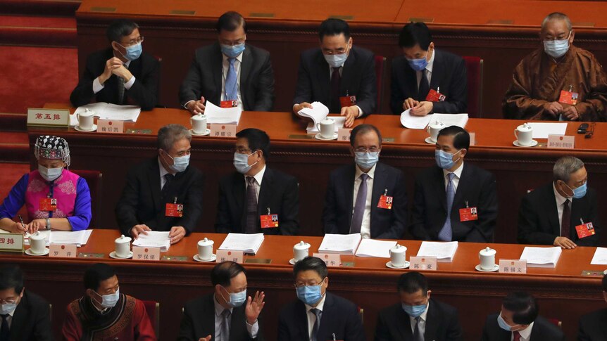 Delegates wear masks and sit spaced out behind desks in a large hall.