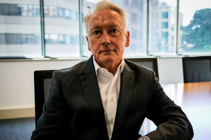 Man wearing suit leans on boardroom table, looking directly into the camera