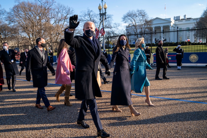 Joe Biden wearing a trench coat and gloves, waving as he walks outside the White House with his family.
