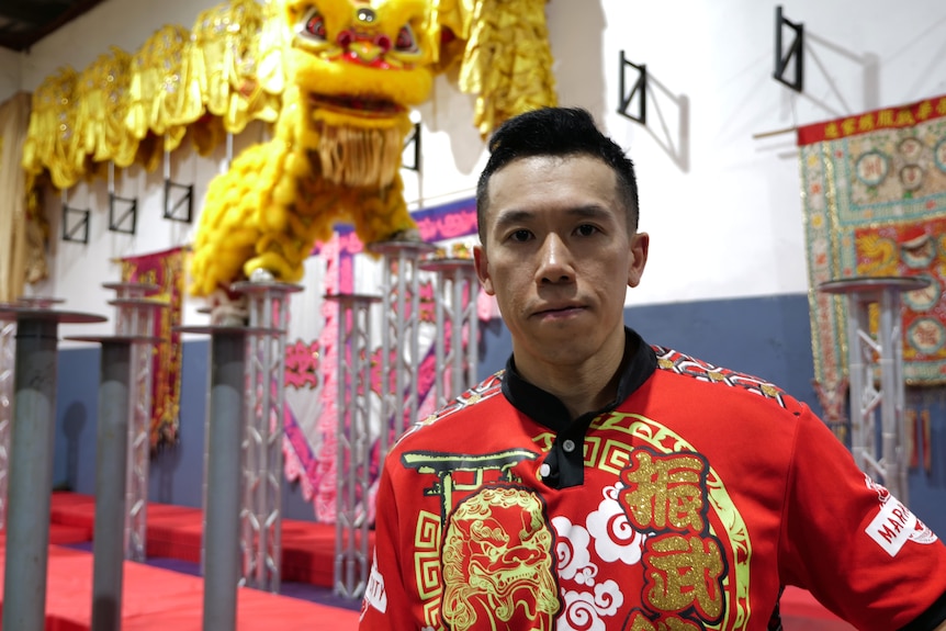 一个身穿印有中国字的红色 T 恤的男人正盯着镜头。 在他身后站着一件黄色的中国狮子服装