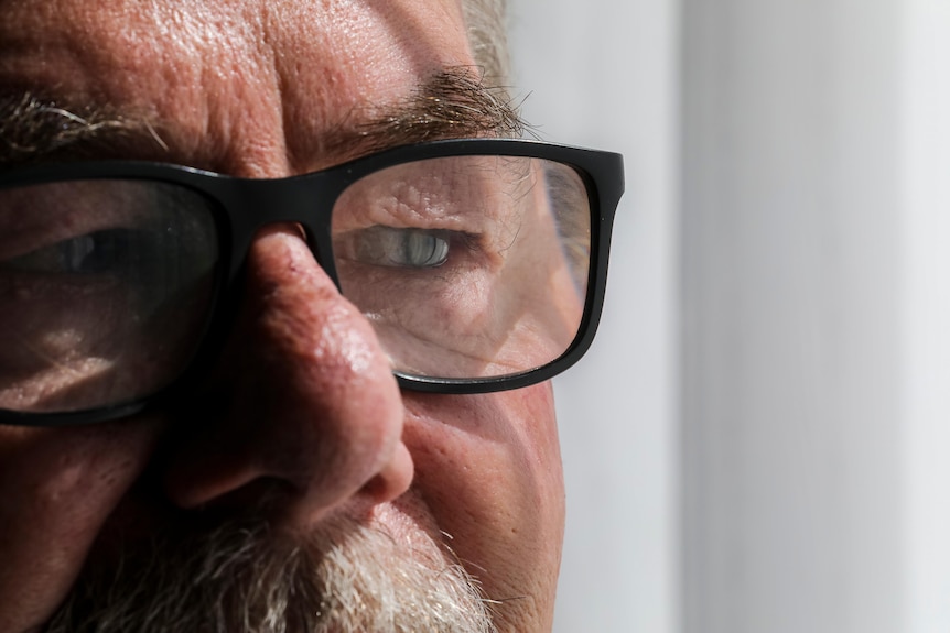 A close-up of a man wearing glasses by a window with tears in his eyes
