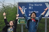 Nell Williams, Viveik Kalra and Aaron Phagura jumping and smiling with arms raised to sky in front of road sign.