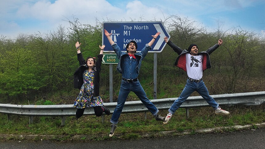 Nell Williams, Viveik Kalra and Aaron Phagura jumping and smiling with arms raised to sky in front of road sign.
