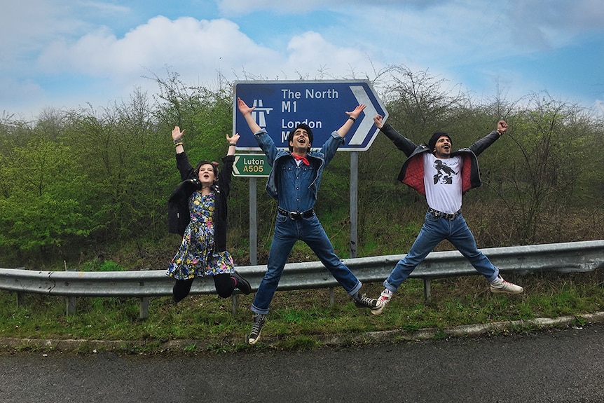 Nell Williams, Viveik Kalra and Aaron Phagura jumping and smiling with arms raised to sky in front of road sign.