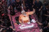 A mam in an orange outfit waves from a car while covered in pink rose petals