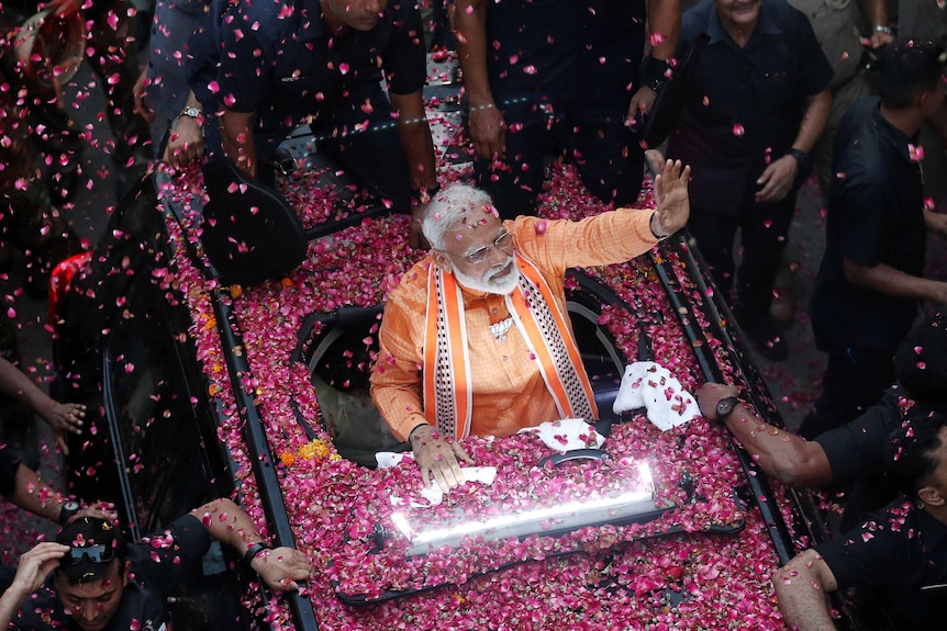 A mam in an orange outfit waves from a car while covered in pink rose petals