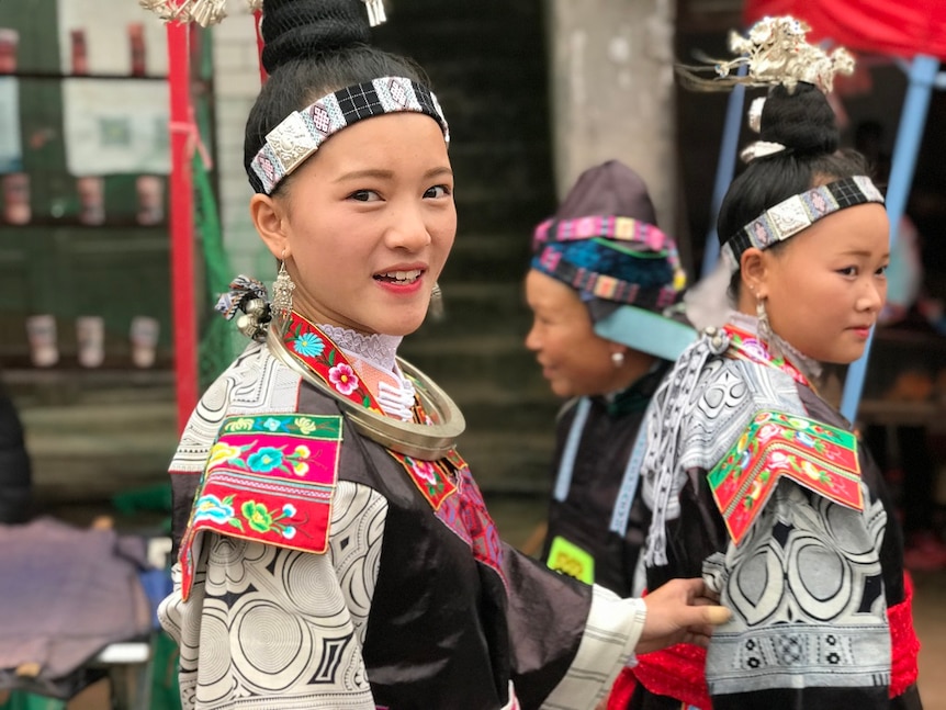 Two women wearing colorful ceremonial coats, jewelery and headwear walk through a rural village.