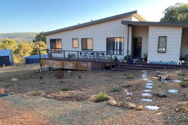 A white weatherboard house with a sloping roof, on a slight hill in a rural valley with a gravel garden out the front.