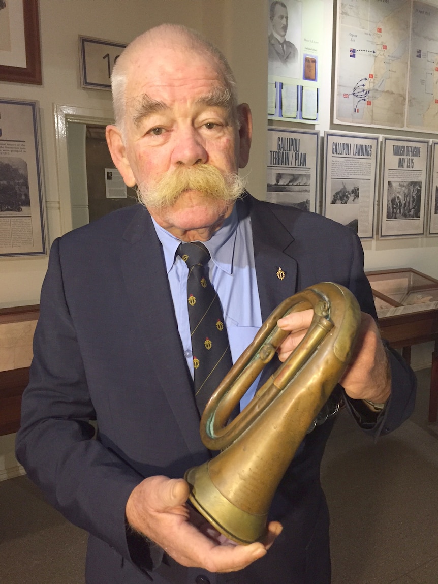 A man holds a bugle. There is military paraphernalia on the wall behind him.