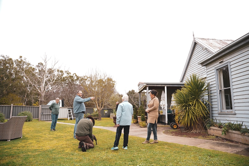 Five men and women stand around a mowed grass area pointing towards a weatherboard building.