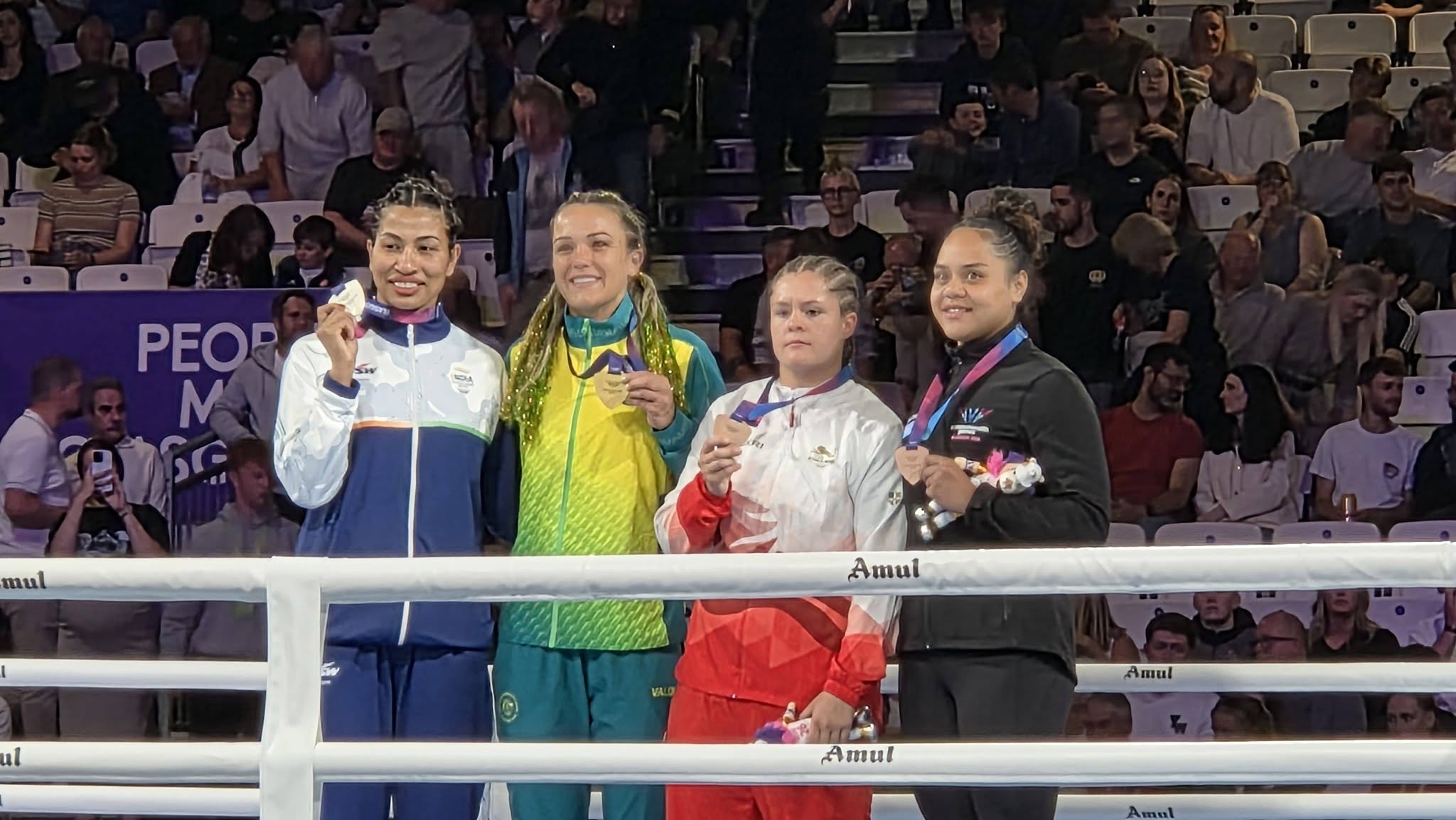 Four young women in different coloured track suits hold up medals around their necks in a boxing ring smiling for a photo