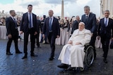 Pope in a all white being wheeled by a man in a dark suit