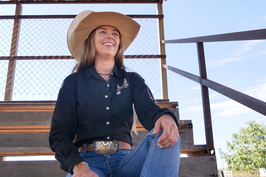 A cowgirl in a navy shirt and brown hat sits in a stand and smiles