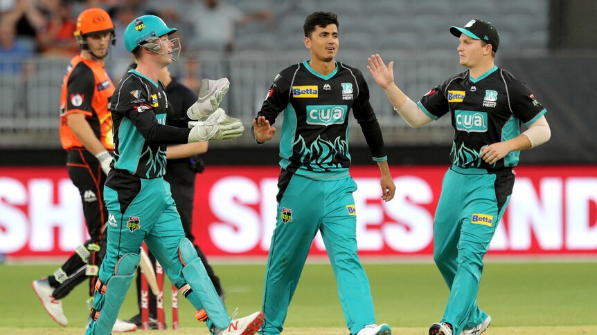 A young south Asian man in green and black cricket uniform is congratulated by teammates in stadium.