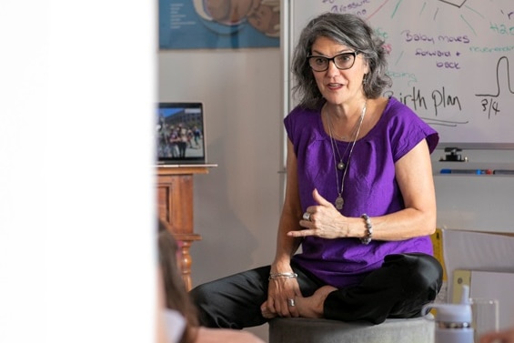 A woman with grey hair and a purple shirt sits in front of a white board talking