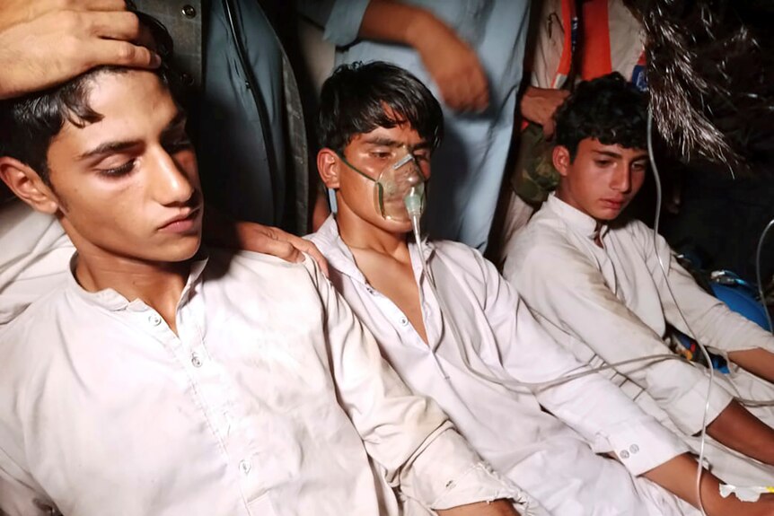 Three teenagers in white shirts sit, with the one in the middle wearing an oxygen mask. 