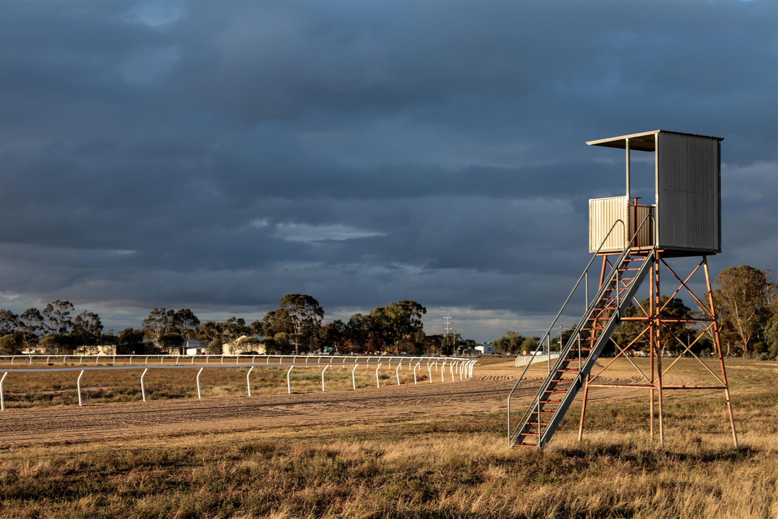 A tin box on a stand with a ladder up to it overlooks a dirt track and white railing of a race track. Clouds and trees behind.