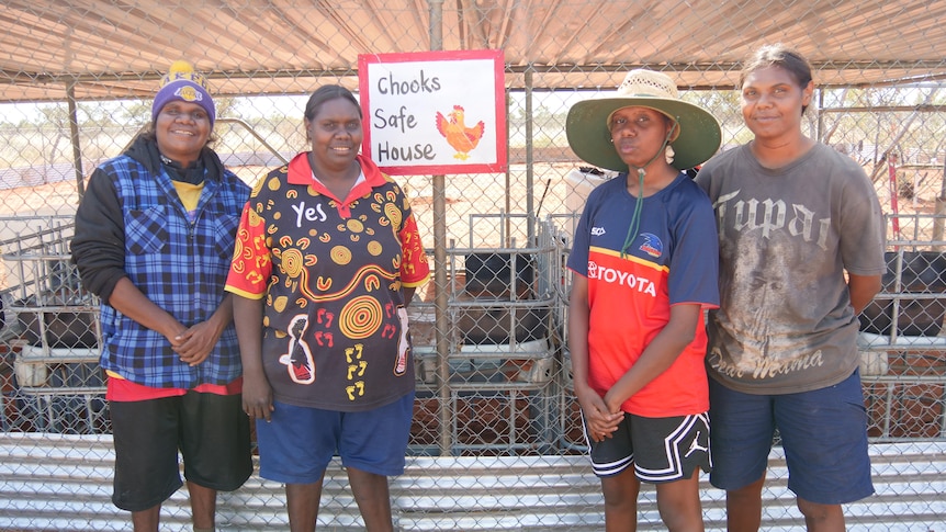 Four Aboriginal women stand two on either side of a sign on their chicken coop which reads 'chooks safe house'.