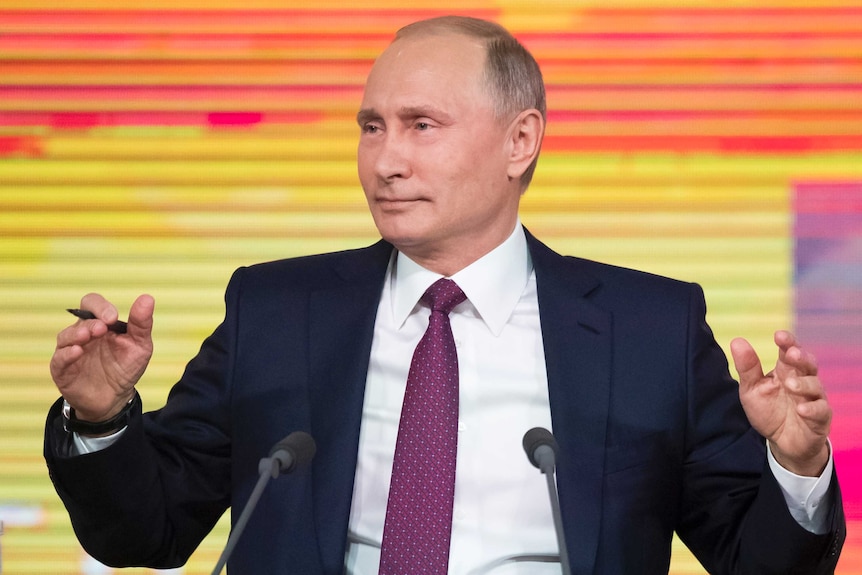 Vladimir Putin gestures as he gives a press conference in front of a colourful screen.