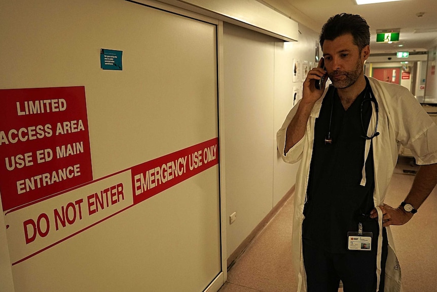A doctor stands in a hospital hallway while he has a phone to his ear.