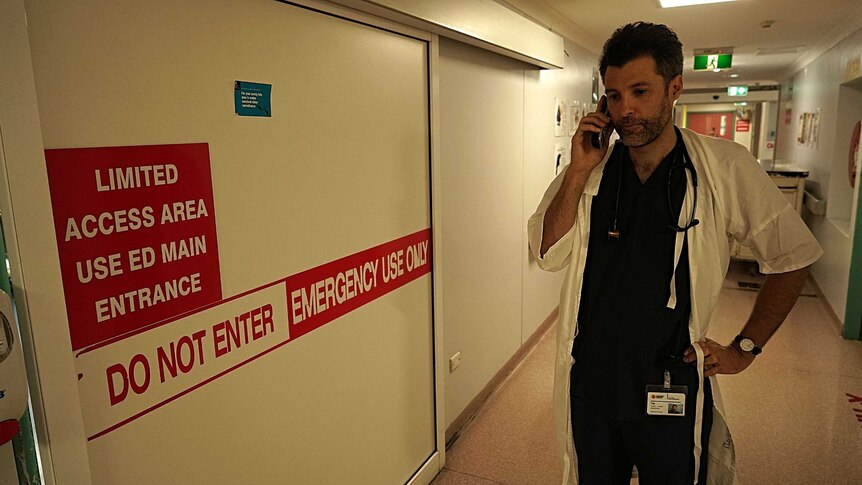 A doctor stands in a hospital hallway while he has a phone to his ear.