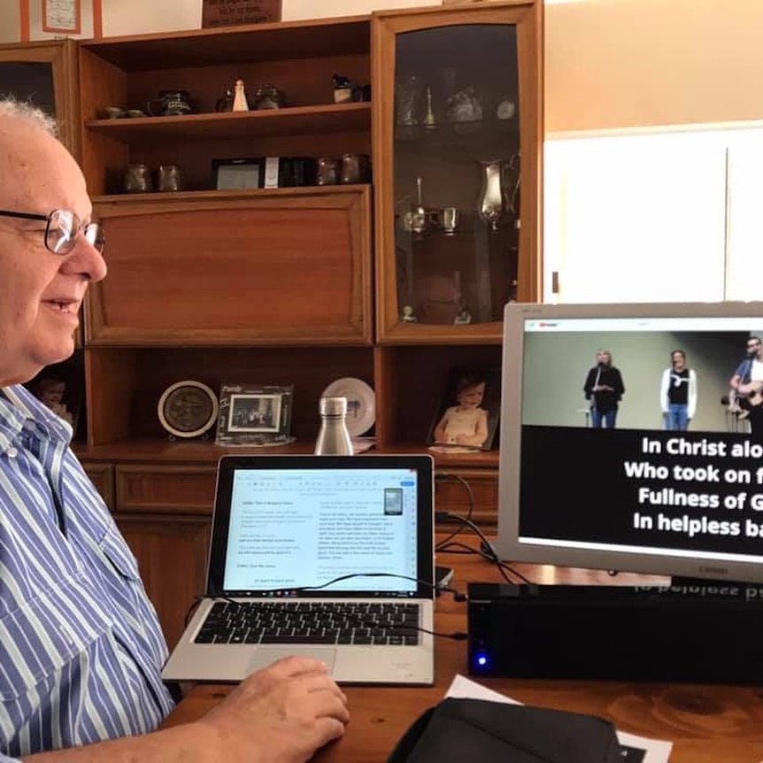 a man sits at a table and watches  church service on his computer