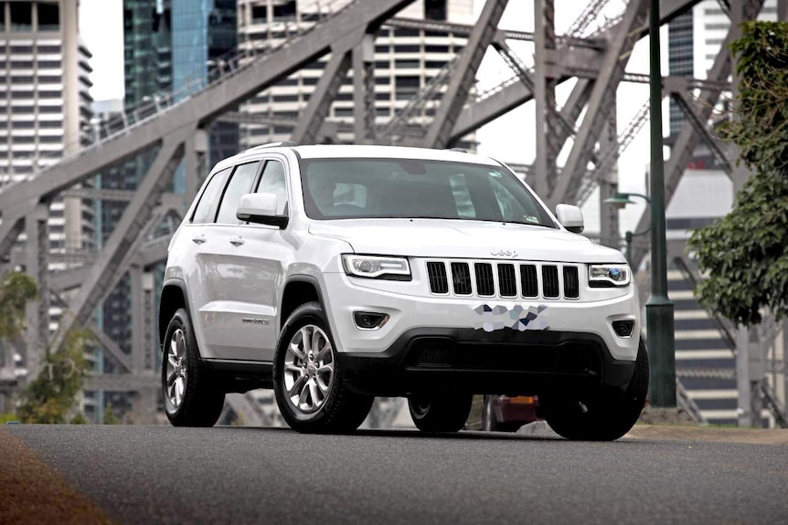 Marketing image of a slick white Jeep SUV parked on the road with a bridge and a cityscape in the background.