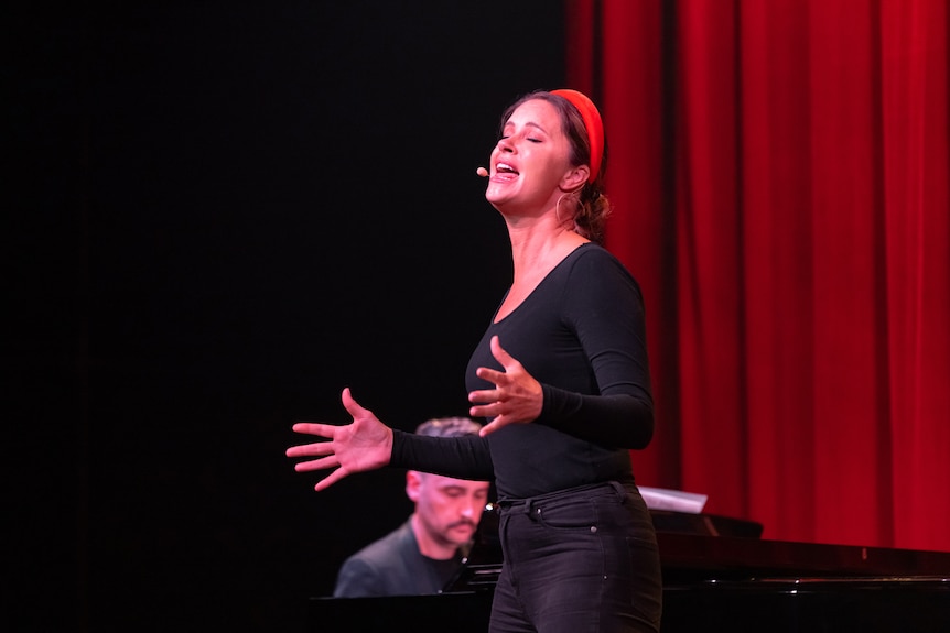 A woman in her early 40s in drama blacks sings, her hands flexed for emphasis, a man playing piano behind her