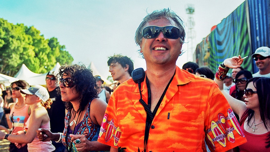 Ken West is outdoors in a crowd, smiling. He wears sunglasses, an orange shirt and a walkie talkie.