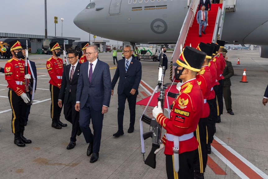 El primer ministro Anthony Albanese está flanqueado por soldados vestidos de rojo mientras camina sobre la pista del aeropuerto con un avión detrás de él.