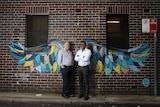 Two men stand in front of colourful street art wings painted on a brick wall