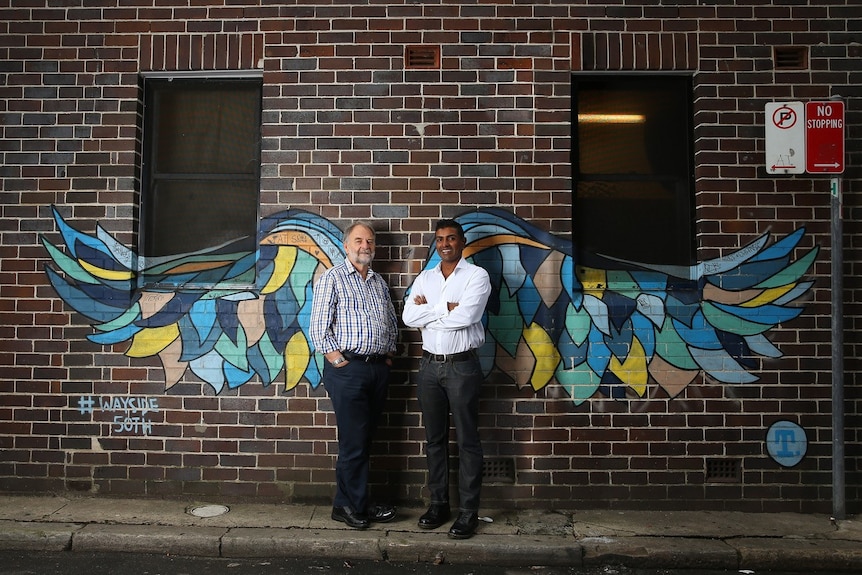 Two men stand in front of colourful street art wings painted on a brick wall