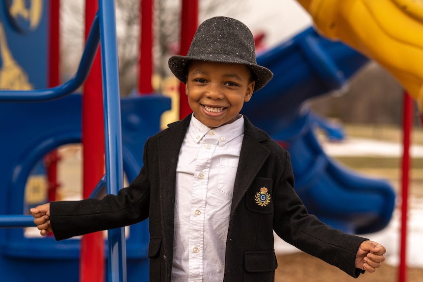 A little African American boy in a grey fedora and blazer, with his arms outstretched at a playground 