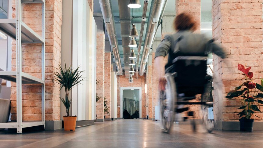 A woman using a wheelchair speeds along a corridor in an industrial building. She is wearing a blazer.