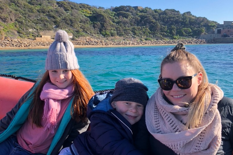 A mother wearing sunglasses and a jumper smiles as she sits in a small boat with her young son and daughter