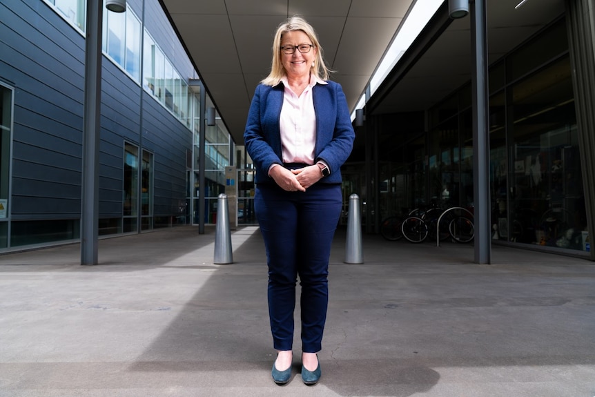 A woman with blonde hair in a navy pant suit and pink shirt stands in front of a modern glass council building