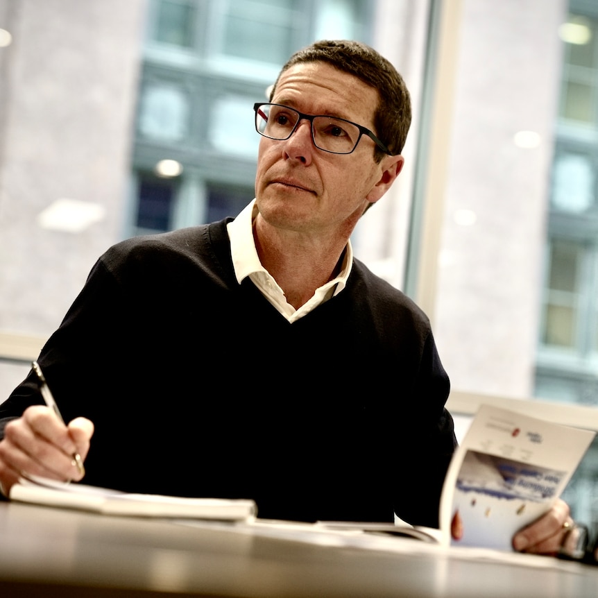 a man sits at his desk with a pen in his hand, he wears glasses and has short brown hair