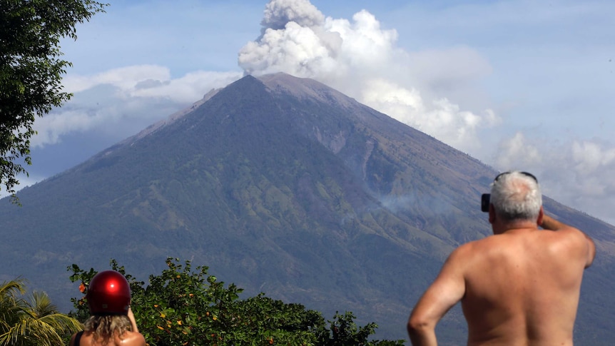 Two tourists watch Mount Agung erupt ash and smoke on a sunny day in Bali.