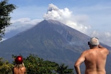 Two tourists watch Mount Agung erupt ash and smoke on a sunny day in Bali.