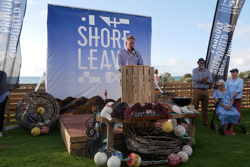Man stands on platform made of pallets, fishing buoys sit in front a sign that says Shore Leave is behind. Sky is blue. 