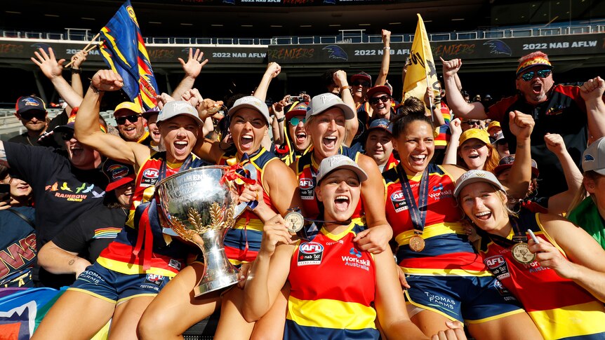 A group of AFLW teammates shout and cheer and pump their fists as they hold the AFLW trophy with fans behind them.