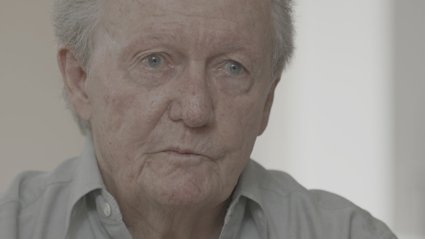 An older man with blue eyes and grey hair talking to someone sitting opposite.