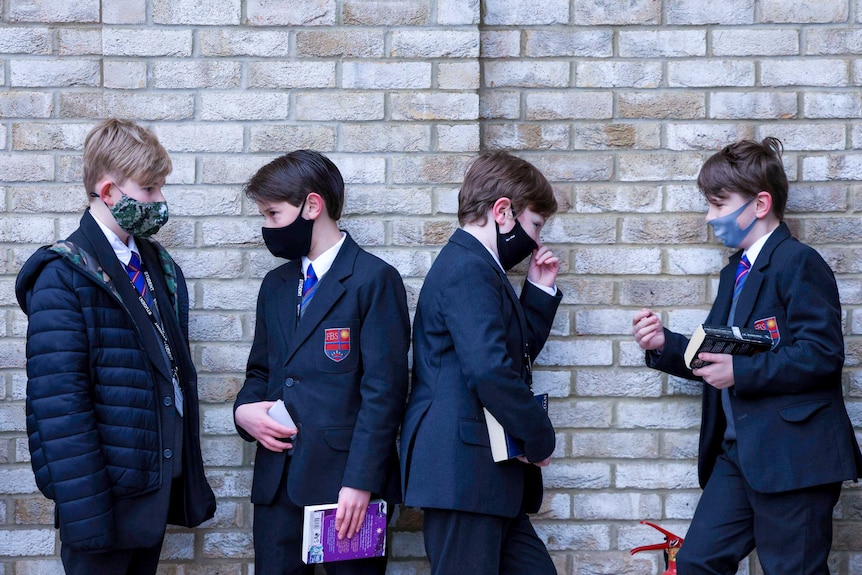 Four boys in blazers and face masks leaning against a brick wall