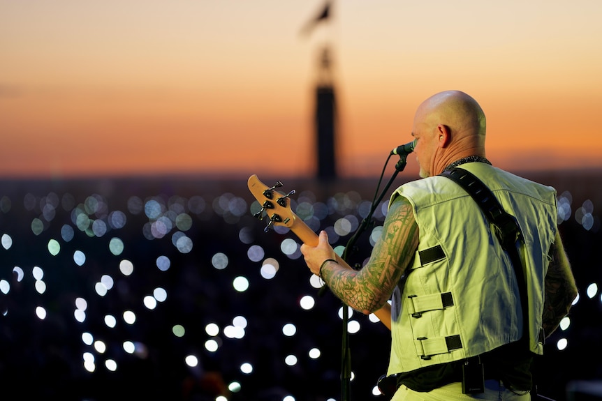 Лысый белый мужчина с татуировкой на рукаве и гитарой в белой куртке играет перед толпой на закате.