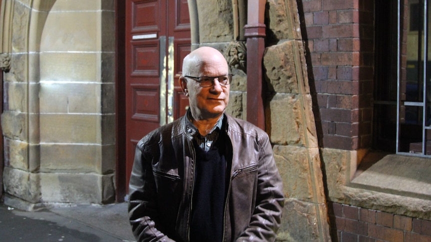 A bald, bespectacled man in a leather jacket stands outside a sandstone and brick building.