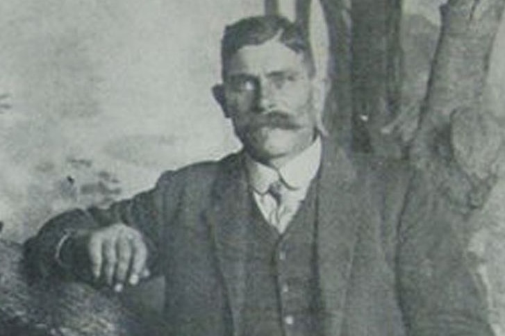 An historic black and white photo of a young man in a suit leaning against a tree.