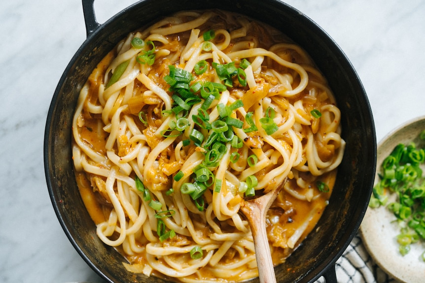 Shallow dutch oven with udon noodles in a kimchi and cheese sauce, topped with sliced spring onions. A quick vegetarian meal.
