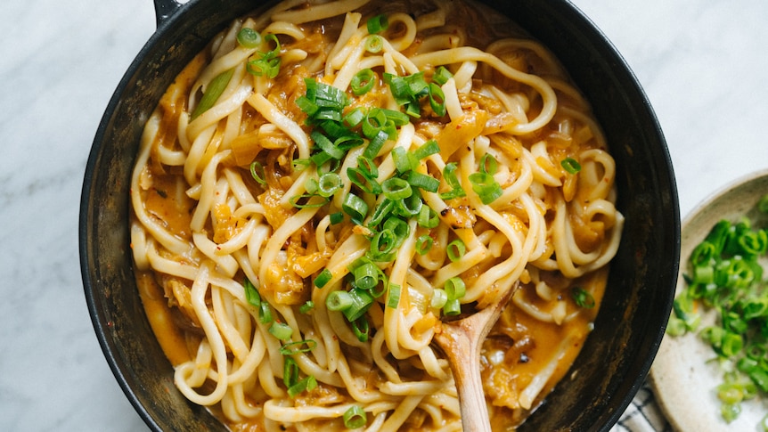 Shallow dutch oven with udon noodles in a kimchi and cheese sauce, topped with sliced spring onions. A quick vegetarian meal.