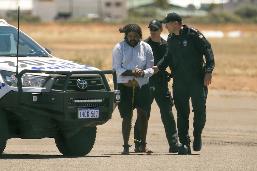 An Indigenous man in shackles in led across a tarmac by two uniformed detectives with a police car next to them.