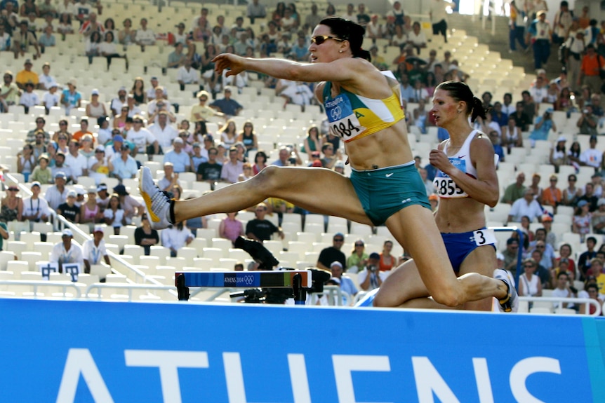 Two women cross hurdles at Olympics event in front of a crowd
